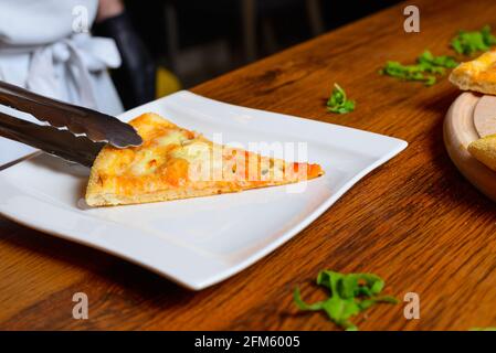 Nahaufnahme des Küchenchefs, der heiße, kitschige Scheiben Pizza mit Ofenzange auf einen quadratischen weißen Teller legt. Stockfoto