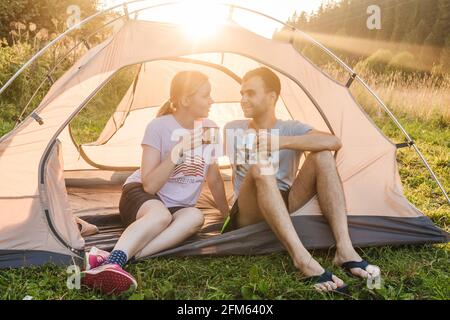 Junge liebende Paar trinken Tee und lächeln im Zelt, Camping in der Natur. Die Sonne geht auf dem Hintergrund auf. Selektiver Fokus. Stockfoto