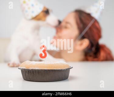 Kaukasische Frau und Jack russell Terrier in Urlaubsmützen betrachten den Kuchen mit einer Kerze. Der Hund und der Besitzer feiern den dritten Geburtstag. Stockfoto