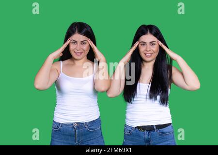 Emotionale Zwillinge in eineiigen Alltagskleidung halten ihren Kopf. Migräne. Kopfschmerzen. Grüner Hintergrund. Stockfoto