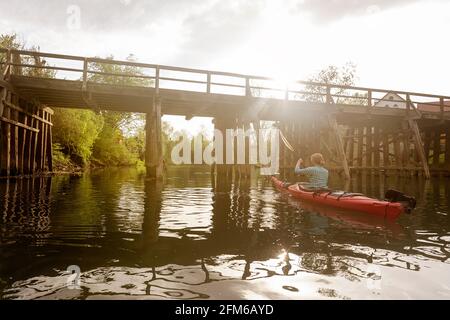 Frau im roten Kajak, die unter einer Holzbrücke über den Fluss Krka in Slowenien mit der umliegenden Kleinstadt Kostanjevica Kajak fährt Stockfoto