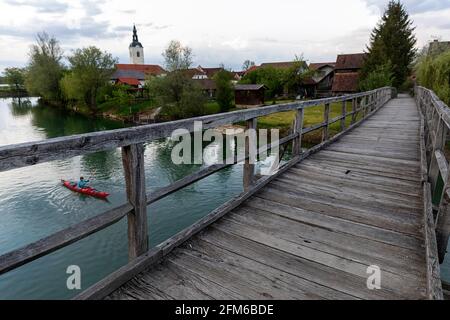 Frau im roten Kajak, die unter einer Holzbrücke über den Fluss Krka in Slowenien mit der umliegenden Kleinstadt Kostanjevica Kajak fährt Stockfoto