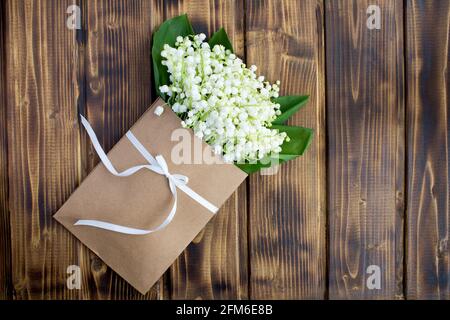 Lilien des Tals in einem Umschlag mit einem grünen Band auf dem braunen Holzhintergrund gebunden.Draufsicht.Copy space.Spring Flowers Konzept. Stockfoto