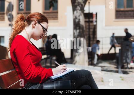 Junge rothaarige Frau mit Brille und in einer roten Bluse sitzend auf einer Bank, während sie etwas in ihr Notizbuch schreibt. Stockfoto