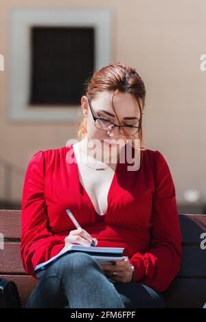 Junge rothaarige Frau mit Brille und in einer roten Bluse sitzend auf einer Bank, während sie etwas in ihr Notizbuch schreibt. Stockfoto