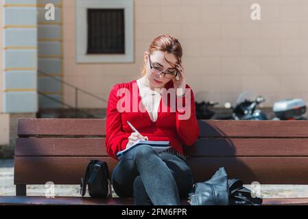 Junge rothaarige Frau mit Brille und in einer roten Bluse sitzend auf einer Bank, während sie etwas in ihr Notizbuch schreibt. Stockfoto