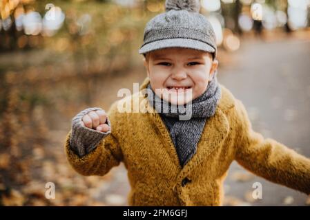 Stilvoller Junge, der Spaß im herbstlichen Stadtpark hat. Glückliches Kind, das zwischen heruntergefallenen Blättern herumläuft. Kindermode. Junge trägt trendigen gelben Mantel, Mütze und Schal. Lächelnder kleiner Junge im Freien. Kinder springen und laufen Stockfoto