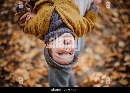 Stilvoller Junge, der Spaß im herbstlichen Stadtpark hat. Glückliches Kind, das zwischen heruntergefallenen Blättern herumläuft. Kindermode. Junge trägt trendigen gelben Mantel, Mütze und Schal. Lächelnder kleiner Junge im Freien. Kinder springen und laufen Stockfoto