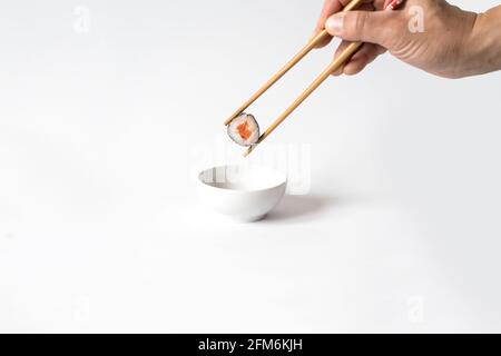 Die Hand des Mannes nimmt Sushi mit Essstäbchen auf und taucht in Sojasauce auf weißem Hintergrund. Stockfoto