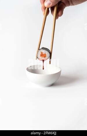 Die Hand des Mannes nimmt Sushi mit Essstäbchen auf und taucht in Sojasauce auf weißem Hintergrund. Stockfoto