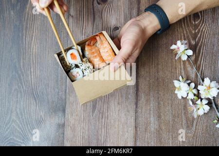 Draufsicht auf einen Mann, der asiatische Speisen zum Mitnehmen mit Essstäbchen auf einem Holztisch isst. Home Delivery-Konzept. Stockfoto