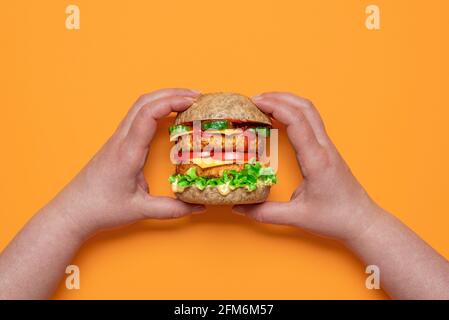 Draufsicht mit Frauenhänden, die einen veganen Cheeseburger halten. Hausgemachter Burger mit pflanzlichen Zutaten isoliert auf einem orangefarbenen Hintergrund Stockfoto