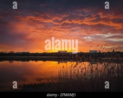 Farbenfroher und dramatischer Sonnenuntergang am Wasser des Teichs. Stille Abendszene am Delia See. Lebhafte Sonnenwolken am Horizont über der Stadt Ungheni Stockfoto