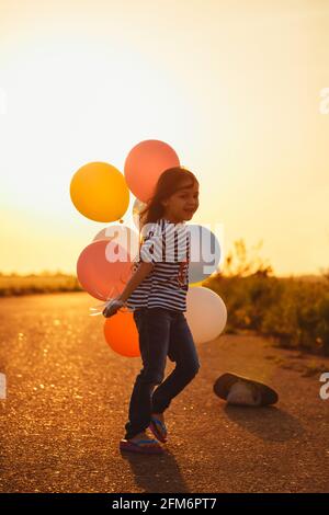 Nettes glückliches kleines Mädchen, das bei Sonnenuntergang mit bunten Luftballons spielt. Selektiver Fokus Stockfoto