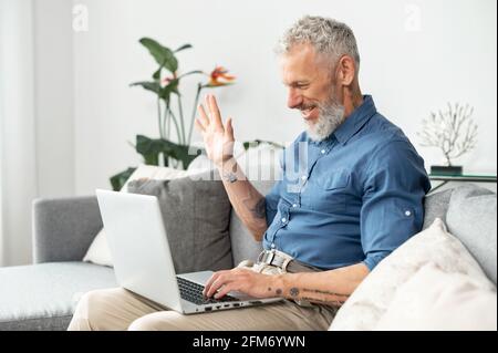 Ein moderner, fröhlicher älterer Mann, der einen Laptop für die Videoverbindung verwendet, ein reifer, grauhaariger Geschäftsmann, der ein legeres Hemd trägt, das in die Webcam winkt und zu Hause auf der bequemen Couch sitzt Stockfoto