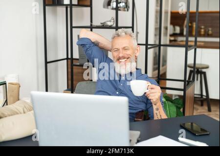 Ein fröhlicher, reifer männlicher Mitarbeiter genießt am Arbeitsplatz im Büro einen Morgenkaffee, sitzt in entspannter Pose mit den Füßen auf dem Tisch, ein zufriedener Mann mittleren Alters in eleganter Freizeitkleidung macht eine Pause Stockfoto