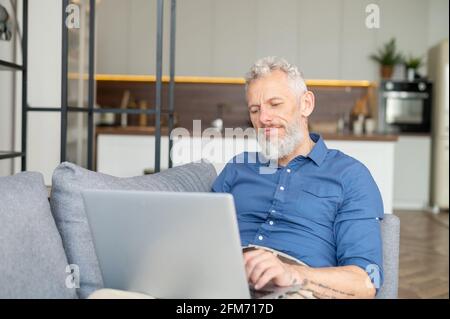 Bärtiger Mann mittleren Alters, der sich mit einem Laptop auf der Couch niederlegt, fernab von zu Hause arbeitet, charismatischer älterer Mann mit lässigem Hemdtippen, E-Mails sendet und in entspannter Atmosphäre zu Hause Nachrichten sendet Stockfoto