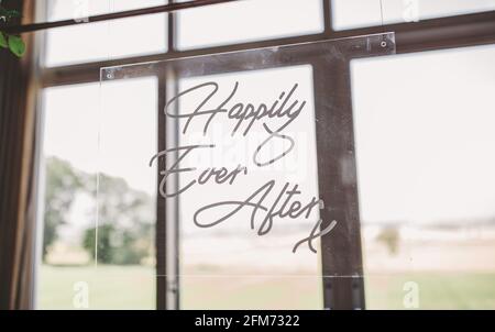 Ein klares Plexiglas-Hochzeitsschild, das glücklich sagt, je nachdem mit Ein Fenster hinter einem Garten Stockfoto