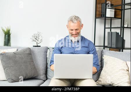 Ein moderner bärtiger Mann mittleren Alters, der einen Laptop benutzt, der zu Hause auf der Couch sitzt. Reifer grauhaariger Mann, der auf der Tastatur tippt, von zu Hause aus arbeitet und in sozialen Medien Nachrichten ausgibt Stockfoto