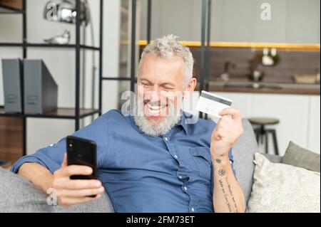 Aufgeregter älterer bärtiger Mann mit Casual Shirt sieht überrascht und glücklich auf dem Telefonbildschirm, moderner Mann mittleren Alters mit Smartphone hebt Hand in Siegesgeste, gewann im Spiel, Ziel erreichen Stockfoto