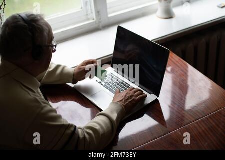 Lächelnder älterer Mann trägt Kopfhörer winken zur Kamera und hat Videoanruf auf dem Laptop, glücklicher älterer Mann mit Kopfhörern sitzt zu Hause auf der Couch und redet mit Modern Stockfoto