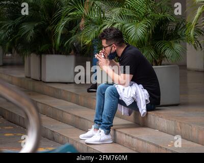 Itagui, Antioquia, Kolumbien - März 7 2021: Ein junger Hispanic-Mann mit einer Maske unter dem Mund sitzt auf einem Schritt und isst und raucht gleichzeitig Stockfoto