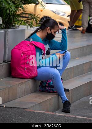 Itagui, Antioquia, Kolumbien - März 7 2021: Junge Hispanic-Frau mit Maske sitzt auf einer Treppe und plaudert am Telefon Stockfoto