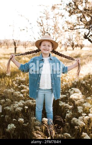 Laghing Kind Mädchen 4-5 Jahre alt mit blonden Zöpfen tragen lässige stilvolle Denim-Kleidung Spaß auf der Wiese im Freien. Kindheit. Frühjahrssaison. Suchen Stockfoto