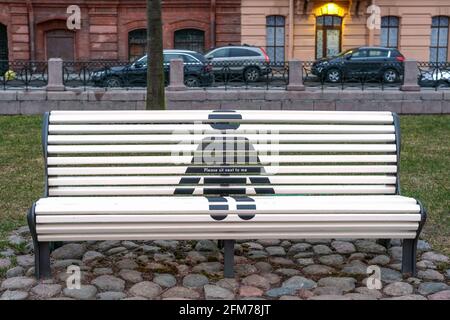 Leere weiße Chatbank in einem öffentlichen Park mit einer speziellen Markierung, wo es sicher ist zu sitzen, Holzsitz, der soziale Distanz hält, neue Pandemievorschriften in freundlicher Form als Menschenfigur Stockfoto