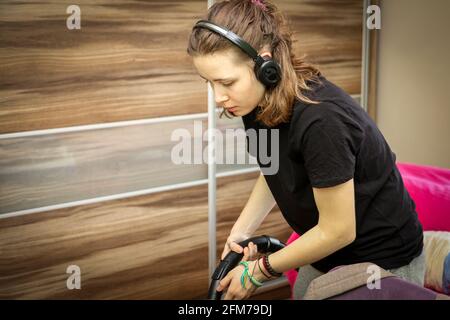 Lässige Moderne Teenage Girl Mit Kabellosen Kopfhörern Reinigungszimmer Mit Staubsauger. Stockfoto