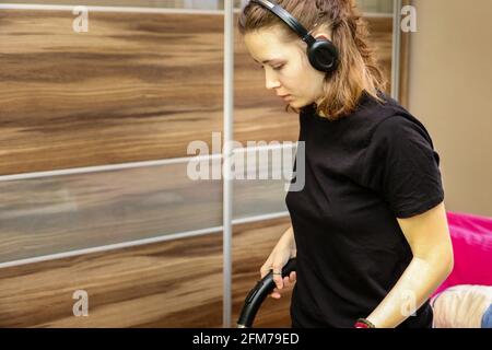 Lässige Moderne Teenage Girl Mit Kabellosen Kopfhörern Reinigungszimmer Mit Staubsauger. Stockfoto