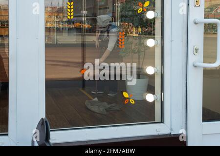 Im Spiegel eines Glasfensters wischt ein Bäckerhandler den Boden mit einem MOP, desinfiziert und reinigt das Geschäft, um es während eines ep sauber zu halten Stockfoto