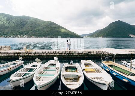 Weiße Boote sind auf dem Dock geparkt. Das Paar umarmt sich vor der Kulisse der Berge und des Meeres. Perast Stadt, Montenegro Stockfoto