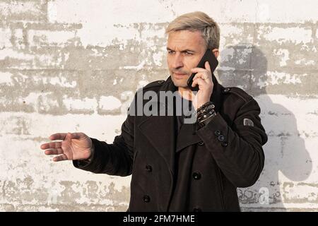 Attraktiver blonder Mann mittleren Alters, der auf einem Handy gegen eine weiße Ziegelwand spricht. Emotionales Gespräch. Männliche Modellierung, Mode-Fotoshooting, prof Stockfoto