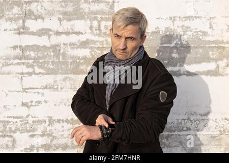 Charmanter blonder Mann mittleren Alters mit Model-Auftritt vor einer weißen Backsteinmauer. Der Mann sieht die Zeit auf seiner Armbanduhr an. Männliches Modell Stockfoto