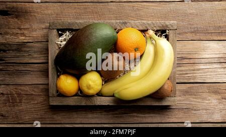 Frische tropische Früchte in einem Holzlieferkarton auf einem Holzhintergrund. Orange, Banane, Mango, Kiwi und Zitrone, flache Lay-Ansicht mit Kopierfläche für Som Stockfoto