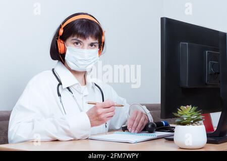 Eine Ärztin, die eine medizinische Schutzmaske trägt, trägt ein Headset, das Sie im Büro ansieht. Der Arzt hört auf die Symptome des Patienten, diagnostiziert Stockfoto