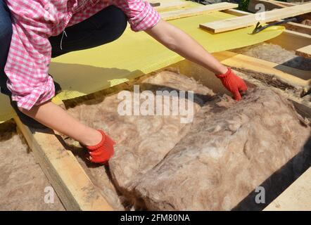 Dachkonstruktion: Eine Heimwerkerin, eine Frau dämmt das Dach mit Mineralwolle, Mineralglasknüppel setzt Dämmmaterial in Deckenverbindungen. Stockfoto