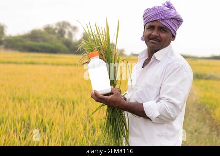 Happy Smiling Farmer hält Reisfeld Ernte und Pestizid oder Chemikalien Düngeflasche in der Hand, indem Sie Kamera in der Nähe von landwirtschaftlichen Ackerland Auf dem Land Stockfoto