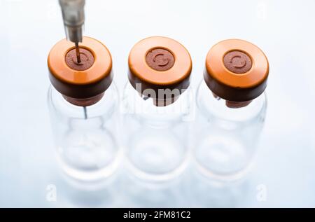 Drei Impfstoffflaschen mit orangefarbenen Kappen in einer Reihe. Eine Spritzennadel klebt durch die rote Gummikappe des linken Fläschchens. Weißer Hintergrund Stockfoto