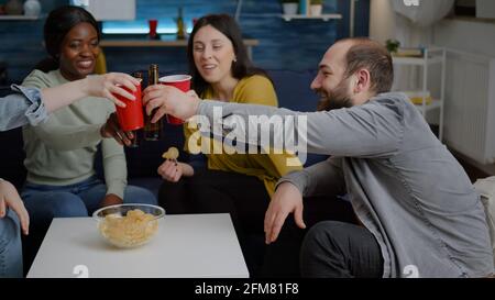 Multikulturelle Freunde geselligen sich, während sie spät in der Nacht auf dem Sofa im Wohnzimmer sitzen und Bier trinken und die Heimparty genießen. Eine Gruppe gemischter Leute, die Zeit miteinander verbringen Stockfoto