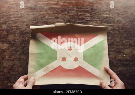 Burundis Nationalflagge auf einem zerrissenen Blatt Papier. Die Hände eines Mannes drücken ein ausgeklapptes Pergamentblatt auf einen braunen Holztisch. Blick von oben Stockfoto