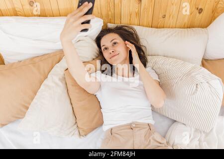 Nahaufnahme einer aufgeregten Frau, die lacht, ein Smartphone hält, ein Selfie macht, Spaß mit einem Gadget hat, auf dem Bett liegt und ihre Freizeit genießt Stockfoto
