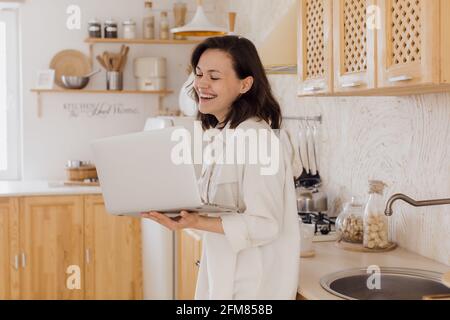 Eine lächelnde junge Frau in einer Küche, genießt ein Videogespräch, lernt sich auf einer Website kennen. Eine Frau verbringt Zeit in einem sozialen Netzwerk Stockfoto