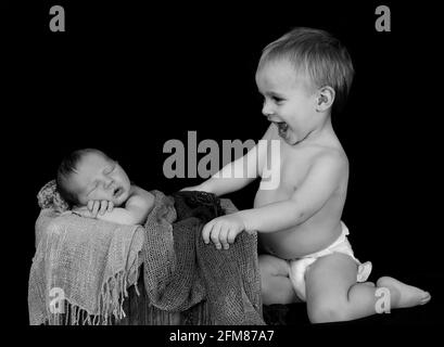 Graustufenaufnahme eines fröhlichen kleinen kaukasischen Jungen mit seinem Neugeborener Bruder auf einem Korb Stockfoto