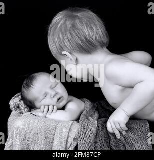 Graustufenaufnahme eines kleinen kaukasischen Jungen mit seinem Neugeborenen Kleiner Bruder auf einem Korb Stockfoto