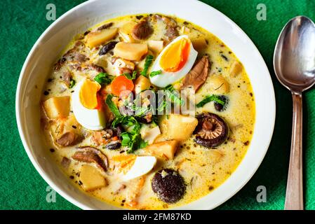 Gesunde, reichhaltige, cremige Suppe mit gekochten Eiern, Kartoffeln, Karotten und Waldpilzen auf weißem Teller, Löffel auf grünem Hintergrund. Stockfoto