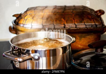 Stahltopf mit kochender sprudelnder Flüssigkeit (Brühe) und aufsteigendem Dampf auf heissem Keramikherd, dahinter alte rustikale Steingutpfanne, Deckel rechts. Stockfoto