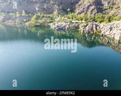 Felsige Ufer des Radon Lake an einem sonnigen Sommermorgen. Luftaufnahme eines alten überfluteten Granitsteinbruchs. Ein malerischer Teich. Stockfoto