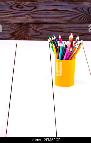 Set aus farbigen Holzstiften in gelbem Kunststoffglas Ein weißer Tisch vor einer Holzwand Stockfoto